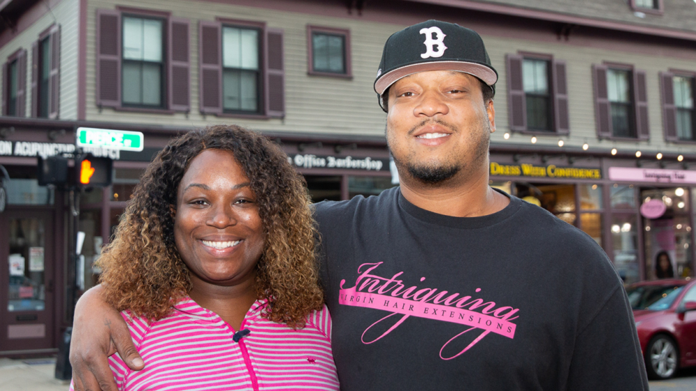 A man and woman smiling with arms around each other, standing in front of a building with shops.